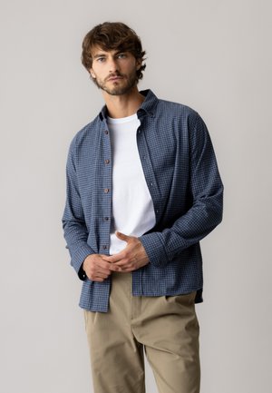 Mand med kort brunt hår iført en blå ternet skjorte over en hvid t-shirt og beige bukser mod en ensartet lys baggrund.