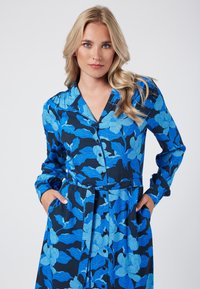 Niebieska sukienka w kwiaty z długimi rękawami, z przodu zapinana na guziki, z kieszeniami. Tkanina ma ciemnoniebieskie tło z jasnoniebieskimi wzorami kwiatowymi.