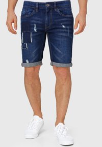 Dunkelblaue Jeansshorts mit abgenutzten Stellen und aufgerollten Säumen. Verfügt über klassischen Fünf-Taschen-Design und Reißverschluss. Getragen mit weißen Turnschuhen.