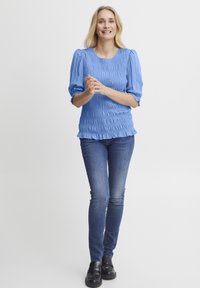Blå, smocket bluse med puffærmer, parret med tætsiddende denimjeans. Stof er letvægts med et tekstureret mønster og flæsekanter.