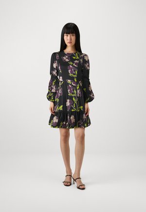 Zwarte jurk met een bloemenprint in paars en groen. Voorzien van opgeblazen mouwen en een gerimpelde zoom. Model draagt zwarte sandalen met bandjes.