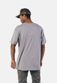 Šedé bavlněné tričko s volným střihem a krátkými rukávy. Zadní strana je hladká, s kulatým výstřihem. Na hlavě je zeleno-beige kšiltovka.