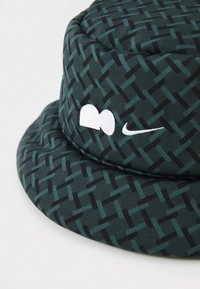 Vihreäkuvioinen sangollinen hattu, joka on valmistettu kankaasta, ja siinä on teksturoitu muotoilu, mustia yksityiskohtia sekä valkoinen Nike-logopistos.