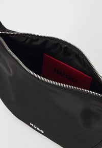 Borsa nera in nylon con chiusura a zip argentata, dotata di una scheda interna rossa con la scritta "HUGO". Branding bianco all'esterno. Texture liscia.