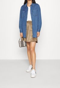 Veste en denim avec deux poches avant, portée sur un T-shirt blanc, accompagnée d'une jupe en daim beige et de baskets blanches. Petit sac métallique.