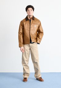 Giacca in finta pelle marrone con colletto marrone, chiusura con zip e tasca laterale; abbinata a pantaloni beige e sneakers marroni su uno sfondo blu.
