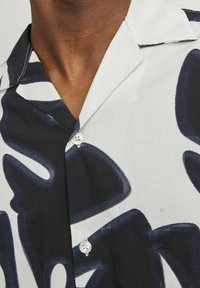 Camicetta a maniche corte con un motivo astratto in bianco e nero, realizzata in tessuto leggero. Presenta un colletto a scollatura e un delicato dettaglio con bottoni.