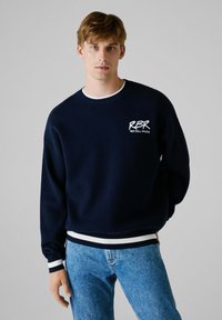 Granatowy sweatshirt z białym logo, żebrowanym dekoltem oraz kontrastującym białym paskiem przy mankietach i dolnym wykończeniu. Noszony z jasnoniebieskimi dżinsami.