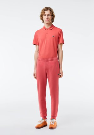 Polo rouge avec col, manches courtes ; joggers assortis rouges avec taille élastique ; modèle portant des baskets oranges avec des lacets blancs.