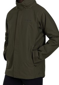 En person som bär en grön vattentät jacka står med händerna i fickorna och visar jackans design och praktiska egenskaper.