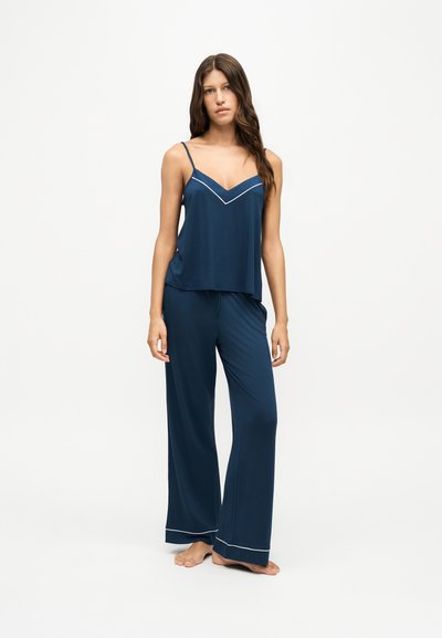 Jeune femme debout, pieds nus, portant un ensemble de pyjama en soie bleu marine avec bordure blanche, comprenant un haut caraco et un pantalon à jambes larges.