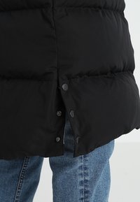 Chaqueta de plumas negra con un diseño de acolchado, que presenta un cierre de botones a presión en el lado izquierdo y una textura suave. Combinada con jeans azules.