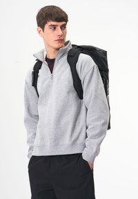 Grau Sweatshirt mit einem Viertel-Reißverschluss, weicher Textur. Schwarzer Rucksack mit sichtbaren Trägern, der modernes Design zeigt. Schlichter Hintergrund.