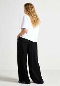 T-shirt bianco a maniche corte indossato con pantaloni neri larghi, che mostrano un design informale e un tessuto testurizzato. Capelli lunghi e mossi parzialmente visibili.