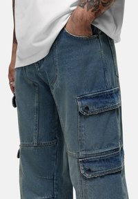 Jasnoniebieskie spodnie cargo z denimu z wieloma kieszeniami, widocznym przeszyciem i luźnym krojem. Kluczowe szczegóły to ciemne zapięcia na guziki oraz kontrastowy materiał.
