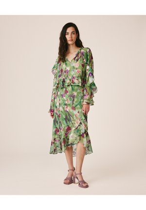 Frau trägt ein grünes Midikleid mit floralem Muster, Rüschenärmeln und -saum, kombiniert mit mauvefarbenen Sandalen mit Blockabsatz, stehend vor einem schlichten Hintergrund.