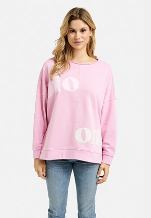 Rosa Sweatshirts mit weißem Text "slo" und "on", lockere Passform, weicher Stoff, relaxter Halsausschnitt und gerippte Bündchen, kombiniert mit blauen Jeans.