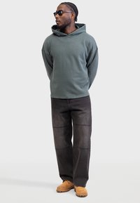 Olijfgroene hoodie, relaxed fit, gecombineerd met zwarte wijde jeans met paneelstiksel en tan suede instappers.