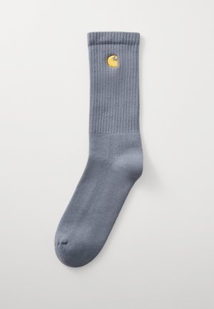 Calcetín de algodón gris con textura acanalada, que presenta un acento de logo circular amarillo cerca de la parte superior. Acabado suave, longitud estándar hasta la pantorrilla.