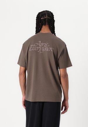 Homme aux cheveux tressés porte un t-shirt marron avec le texte "Air is Everywhere" et un petit logo Nike au dos sur fond blanc.
