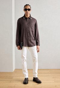 Chemise à manches longues rayée brune avec une poche, associée à un jean blanc et des bottines noires. Le modèle tient une pochette noire et porte des lunettes de soleil.