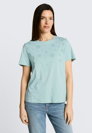 Kobieta ubrana w jasnoniebieską koszulę z krótkim rękawem z fakturowanymi wzorami kwiatowymi i jasnoniebieskie dżinsy, stojąca na jednolitym tle.