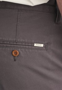 Donkergrijze broeken van soepel materiaal, voorzien van een houten knoop en een GANT-label op de achterzak. Netjes zichtbare stikseldetails.