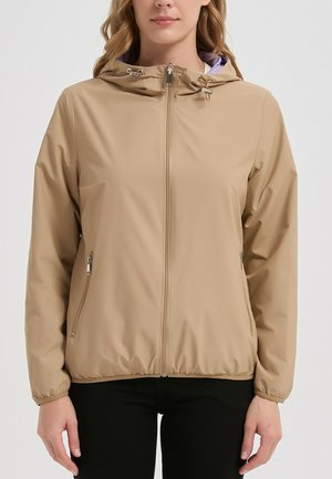 Femme portant une veste à capuche marron clair avec fermeture éclair, cordons et poches latérales zippées, associée à un pantalon noir, sur un fond uni.