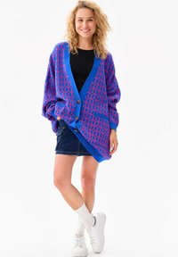 Blauwe cardigan met een roze hexagonaal patroon, oversized pasvorm, twee voorzakken, grote knopen, gecombineerd met een denim rok en witte sneakers.