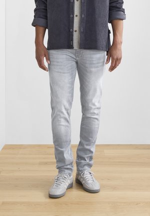Jeans gris claro con corte ajustado y un desvanecimiento sutil, combinados con zapatillas gris. La camisa es de pana en color azul marino con un patrón a cuadros por debajo.