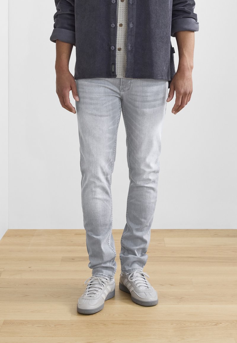 Ljusgrå jeans med slim fit och subtil blekning, kombinerade med grå sneakers. Skjortan är marinblå kordflana med ett rutigt mönster under.