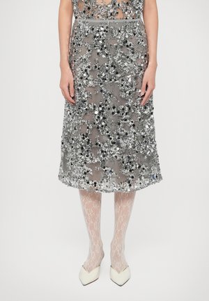 Zarter grauer Midirock, verziert mit schwarzen und silbernen Pailletten, mit leicht ausgestelltem Schnitt, kombiniert mit strukturierten, blassrosa Strumpfhosen und spitzen Schuhen.