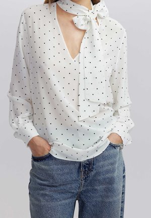 White long-sleeve blouse with black polka dots and a tied neck scarf, paired with blue speckled denim jeans, hands in pockets.