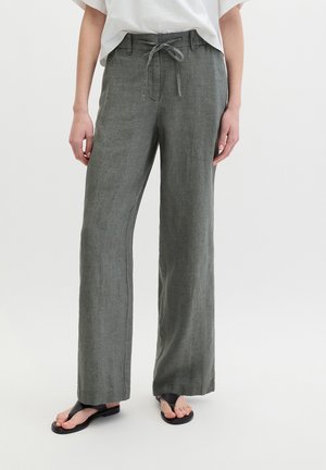 Modell trägt locker sitzende graue Leinenhose mit Kordelzug in der Taille und schwarze Zehensandalen, steht vor einfachem Hintergrund.