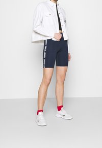 Marina cykelbyxor med vit "TOMMY JEANS"-text på sidan, i kombination med en vit kortärmad knapplöst skjorta och vita sneakers med röda detaljer.