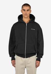 Juodas su zipu megztinis, pagamintas iš medvilnės, su priekinėmis kišenėmis, praktiškomis apykaklėmis ir gobtuvu. Pabrėžtas mažas kontrastingos spalvos logotipas.