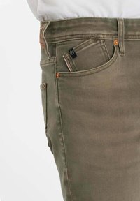 Primo piano di una persona che indossa jeans verde oliva con tasca frontale visibile, taschino portamonete, passante per cintura e rivetti in rame.