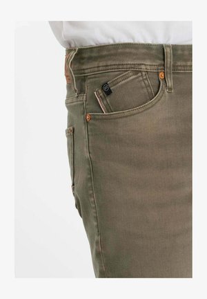 Gros plan d'une personne portant un jean vert olive avec une poche avant visible, une petite poche à monnaie, une passants de ceinture et des rivets en cuivre.
