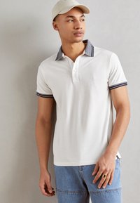 Polo cu mânecă scurtă albă, cu guler și mansete în dungi bleumarin, cu închidere cu butoane și un detaliu subtil al logo-ului. Purtați cu blugi din denim bleu deschis.