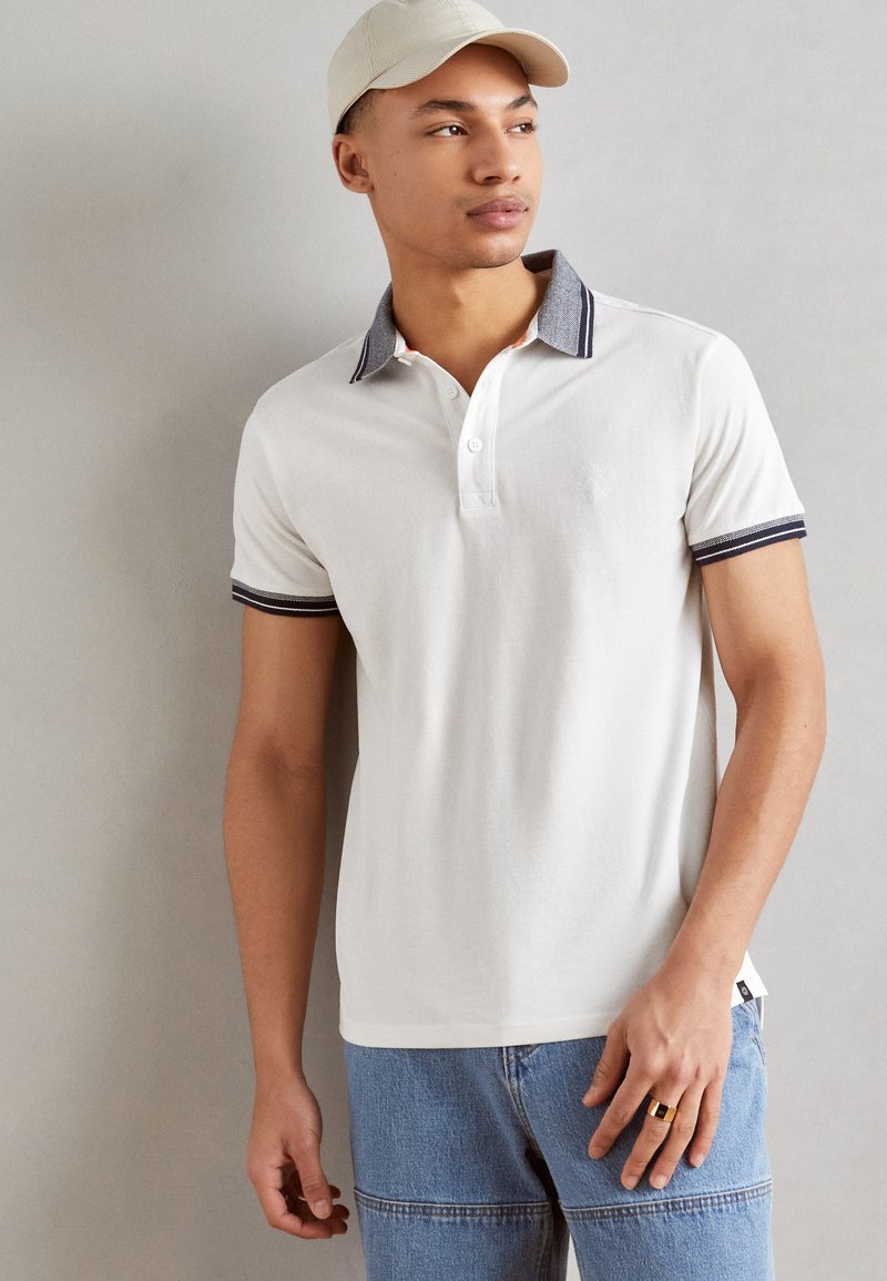 Polo cu mânecă scurtă albă, cu guler și mansete în dungi bleumarin, cu închidere cu butoane și un detaliu subtil al logo-ului. Purtați cu blugi din denim bleu deschis.