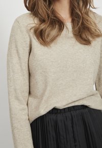 Beige gebreide trui met lange mouwen, ronde hals en subtiele textuur, gecombineerd met een zwarte geplooide rok bij de taille.