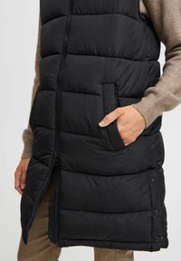 Gilet matelassé noir avec une fermeture éclair à l'avant, des poches latérales, une texture matelassée et des accents de bouton-pression le long de l'ourlet.