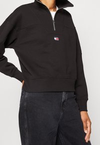 Felpa nera con collo alto, zip a quarto e patch con logo sul petto. Polsini e orlo a coste, realizzata in tessuto morbido.