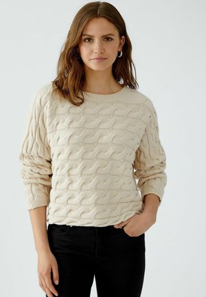 Pull en tricot beige avec un motif en câble texturé, col rond et manches longues, associé à un pantalon noir.
