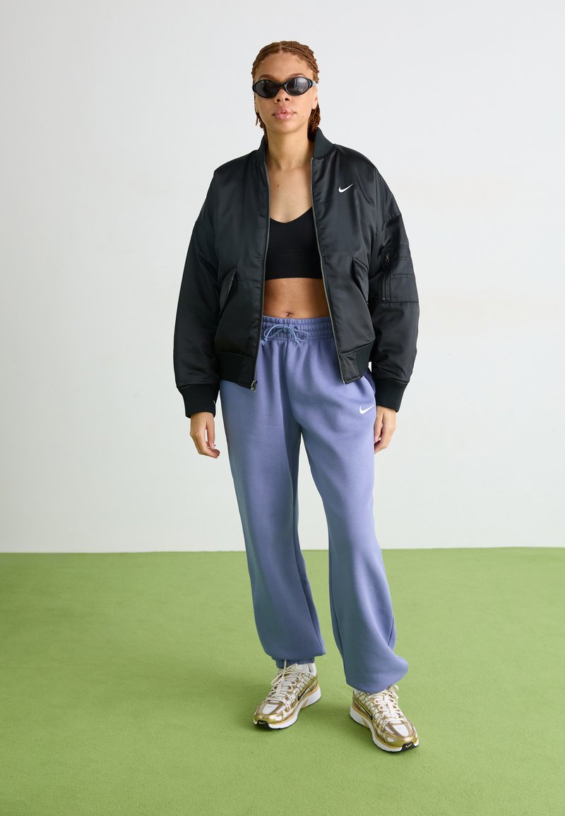 Veste bomber noire, crop top noir, pantalon de jogging violet clair et baskets avec des accents dorés. Les accessoires incluent des lunettes de soleil sombres.