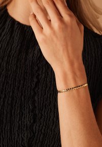 Bracelet en or avec un design de chaîne maillée, porté sur un poignet, sur un haut noir texturé. Accent sur le métal brillant et le motif complexe.