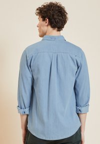 Camicia in denim blu chiaro con vestibilità rilassata, colletto button-down, maniche arrotolate e dettaglio di piega sul retro. Consistenza morbida, design casual.