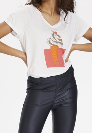 Kobieta ubrana w białą koszulkę z grafiką rożka lodowego oraz czarne spodnie z sztucznej skóry, dłonie opierające się na biodrach.