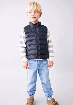 Copil blond purtând o vestă neagră matlasată peste un pulover cu dungi gri și albe, blugi bleu deschis și pantofi bej, stând în fața unui fundal alb.