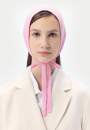 Junge Frau mit hellem Teint und braunen Haaren trägt eine rosa gestrickte Mütze, die unter dem Kinn gebunden ist, und einen weißen Mantel mit Kragen.